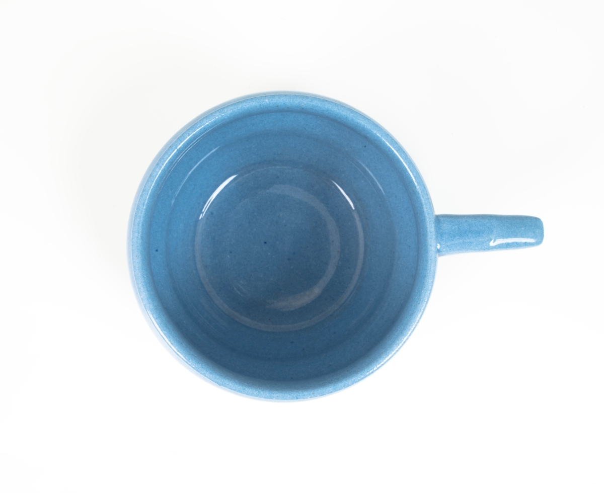 Tekopp med underskål i glasert steingods med begitning. Flattrykket kuleformet korpus, påsatt hank med knekk, samt sirkulær underskål. Kopp og skål er lyseblå og dekorert med en stilisert bord i grønt og rødt, samt horisontale grønne linjer som understreker deres form.