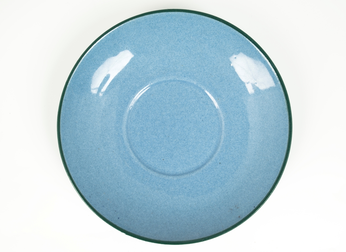 Tekopp med underskål i glasert steingods med begitning. Flattrykket kuleformet korpus, påsatt hank med knekk, samt sirkulær underskål. Kopp og skål er lyseblå og dekorert med en stilisert bord i grønt og rødt, samt horisontale grønne linjer som understreker deres form.