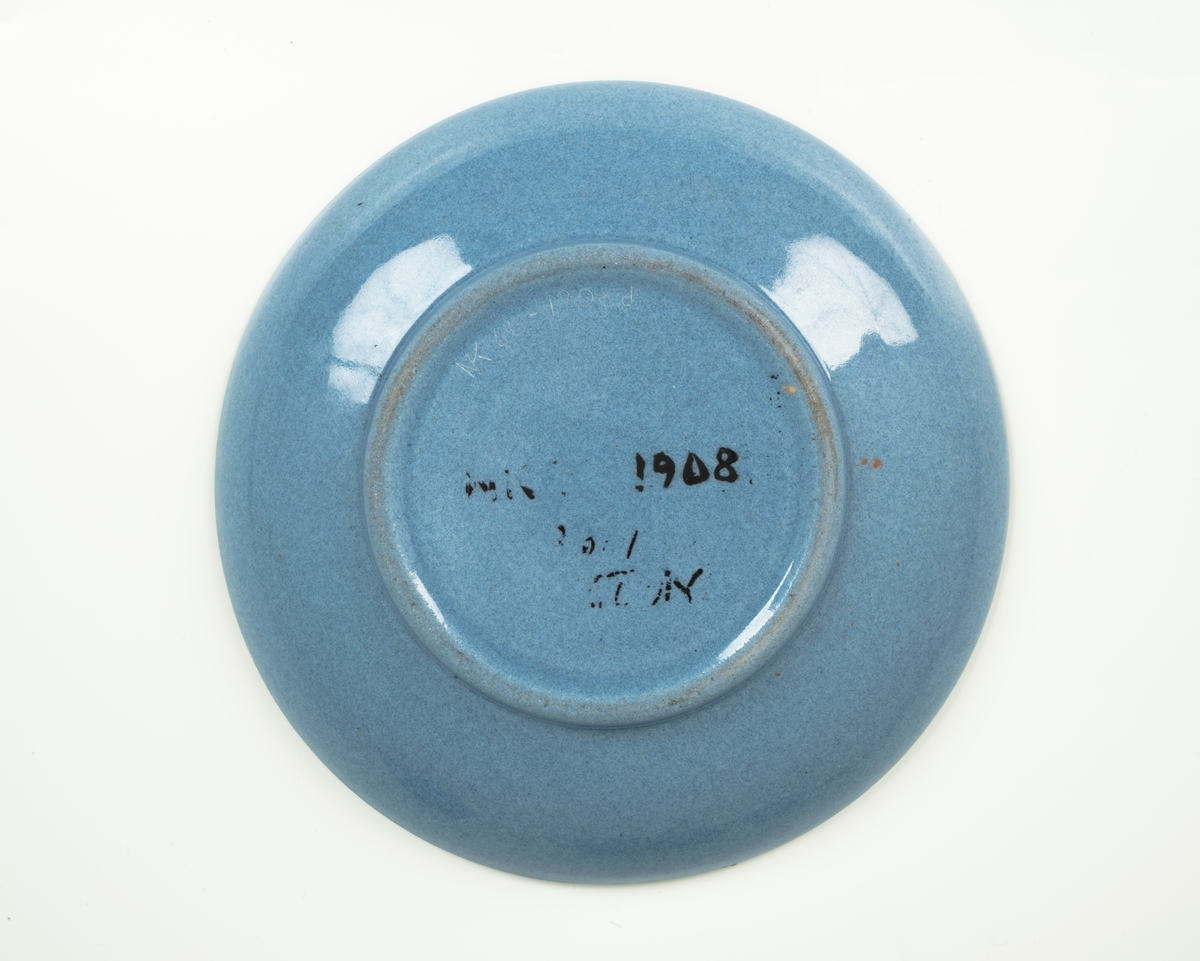 Tekopp med underskål i glasert steingods med begitning. Flattrykket kuleformet korpus, påsatt hank med knekk, samt sirkulær underskål. Kopp og skål er lyseblå og dekorert med en stilisert bord i grønt og rødt, samt horisontale grønne linjer som understreker deres form.