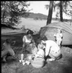 Kvinnor och barn sitter på en filt med fikakorg vid en strand, kaffekoppar och termosar på filten. En eka ligger upp och ned bakom dem. Kvinnan till höger är sannolikt Birgitta Berglund.