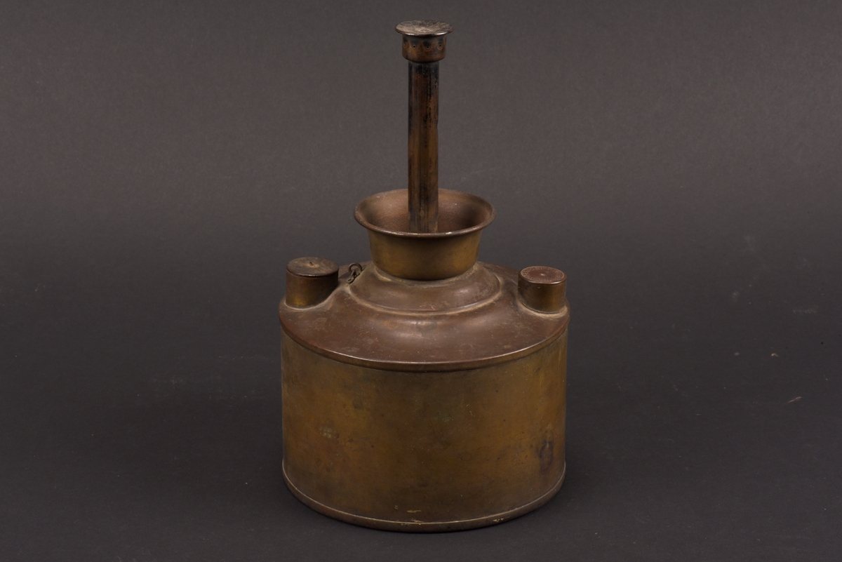 Cylindrisk gasoljelampa tillverkad av mässing. I mitten en cylindrisk vekhållare som är fäst i gängat påfyllnadslock. På sidorna av oljehuset sitter två cylindriska upphöjningar med oklar funktion. Den ena är öppningsbar och locket har ursprungligen varit förankrad i oljehuset via en länk.