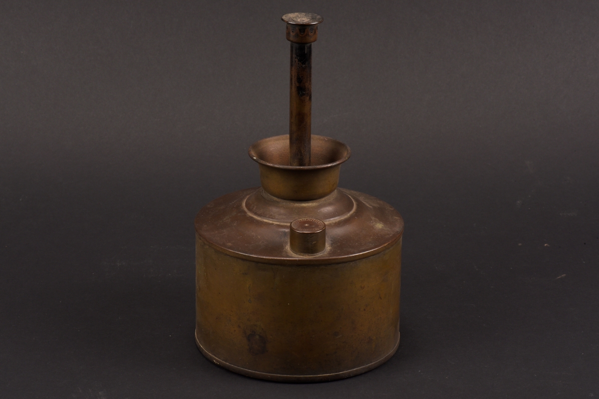 Cylindrisk gasoljelampa tillverkad av mässing. I mitten en cylindrisk vekhållare som är fäst i gängat påfyllnadslock. På sidorna av oljehuset sitter två cylindriska upphöjningar med oklar funktion. Den ena är öppningsbar och locket har ursprungligen varit förankrad i oljehuset via en länk.