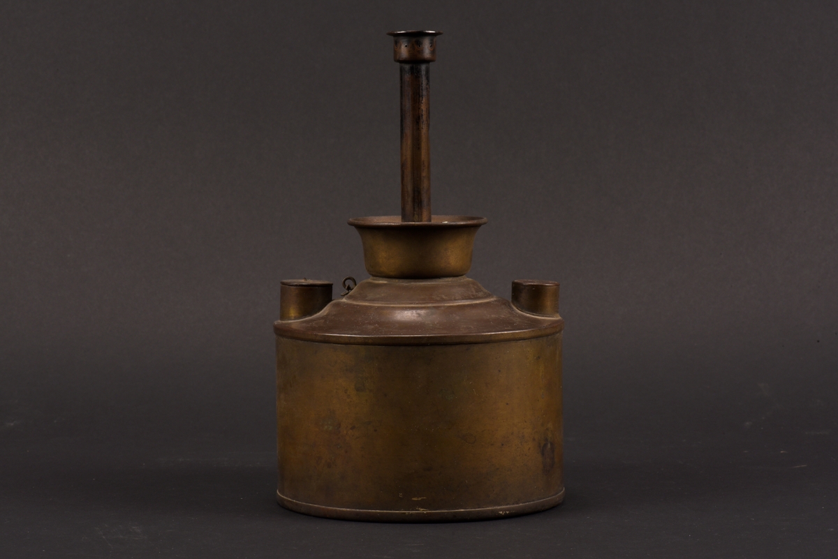 Cylindrisk gasoljelampa tillverkad av mässing. I mitten en cylindrisk vekhållare som är fäst i gängat påfyllnadslock. På sidorna av oljehuset sitter två cylindriska upphöjningar med oklar funktion. Den ena är öppningsbar och locket har ursprungligen varit förankrad i oljehuset via en länk.