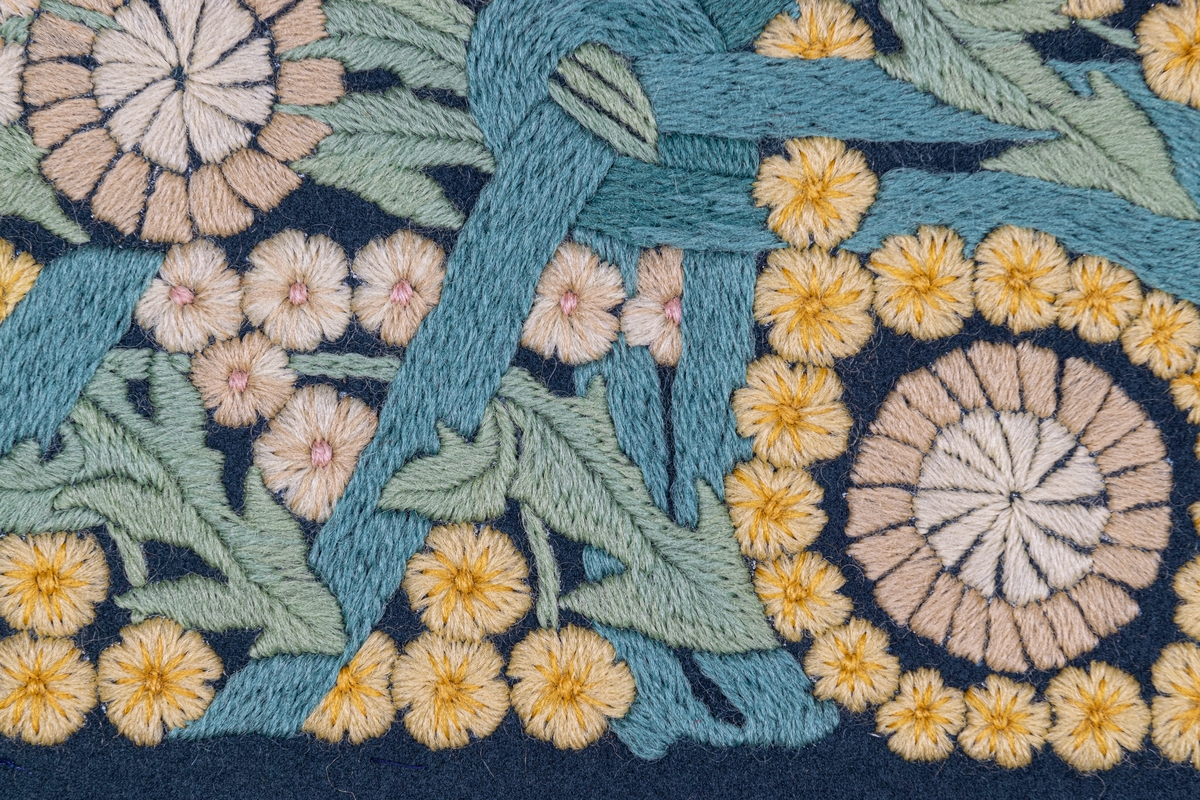 Påbörjad dyna, 80 x 58. Broderi i schattersöm i ullgarn i ljusa färger på blått kläde. Parti ur stor bonad av Ferdinand Boberg. Blommor och bladverk i fält inramat av gullvivor. Ljusgult, vitt, grönt, blått, rött m.fl. färger.