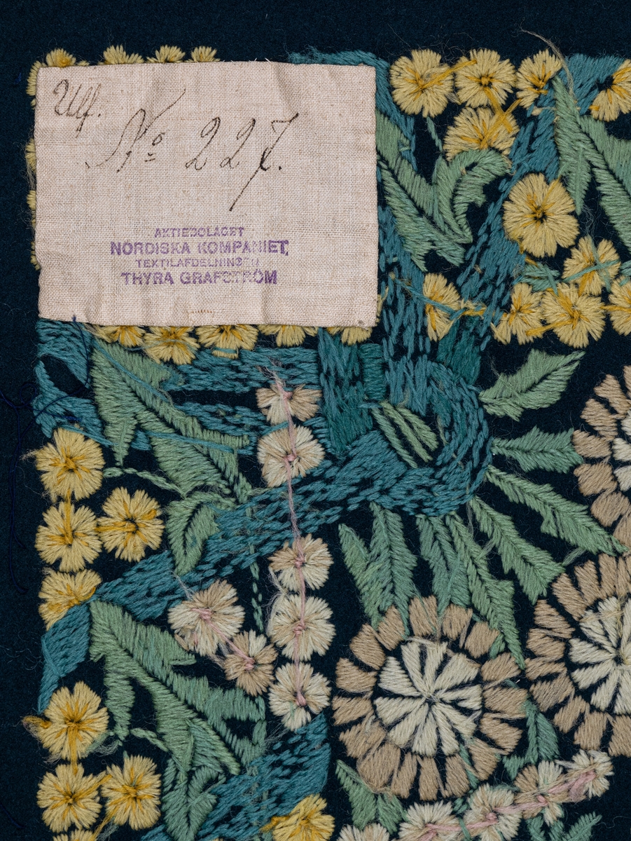 Påbörjad dyna, 80 x 58. Broderi i schattersöm i ullgarn i ljusa färger på blått kläde. Parti ur stor bonad av Ferdinand Boberg. Blommor och bladverk i fält inramat av gullvivor. Ljusgult, vitt, grönt, blått, rött m.fl. färger.