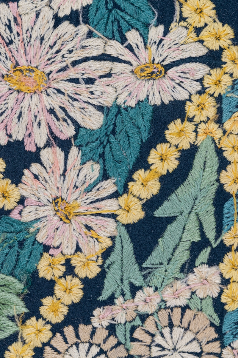 Påbörjad dyna, 80 x 58. Broderi i schattersöm i ullgarn i ljusa färger på blått kläde. Parti ur stor bonad av Ferdinand Boberg. Blommor och bladverk i fält inramat av gullvivor. Ljusgult, vitt, grönt, blått, rött m.fl. färger.