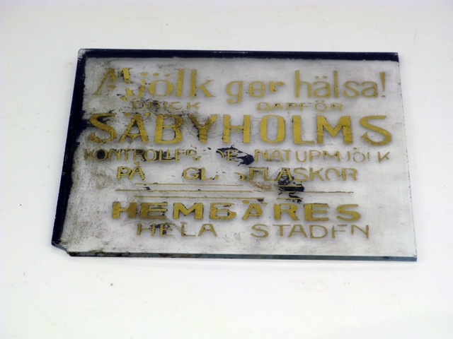 Glasskyltar, 4 st med förgylld text. Bottenfärgen avflagnad på skyltarna.
1) Text: Mjölk ger hälsa, drick därför Säbyholms kontrollerade naturmjölk.
2) Text: Eleganta Automobiler tillhandahålles dygnet om. Tel. 277. A.B. O. Magnusson.
3) Harry Lindberg. Utför alla slags Stenarbeten. Landskrona Kalkbruk, Ständigt lager av kalk.
4) Text: Café Temperance, Frukostar, middagar, supéer.
Inrammad i en svart trätam med krokar upptill för upphängning. Glasskylten har spruckit på två ställen.