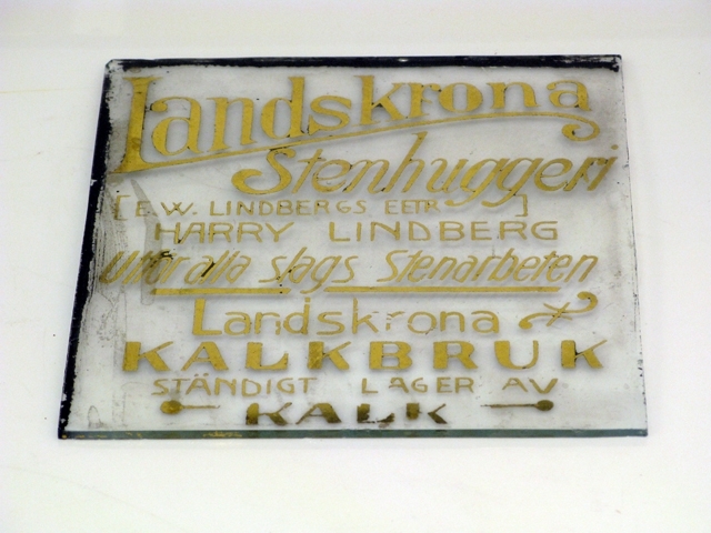 Glasskyltar, 4 st med förgylld text. Bottenfärgen avflagnad på skyltarna.
1) Text: Mjölk ger hälsa, drick därför Säbyholms kontrollerade naturmjölk.
2) Text: Eleganta Automobiler tillhandahålles dygnet om. Tel. 277. A.B. O. Magnusson.
3) Harry Lindberg. Utför alla slags Stenarbeten. Landskrona Kalkbruk, Ständigt lager av kalk.
4) Text: Café Temperance, Frukostar, middagar, supéer.
Inrammad i en svart trätam med krokar upptill för upphängning. Glasskylten har spruckit på två ställen.
