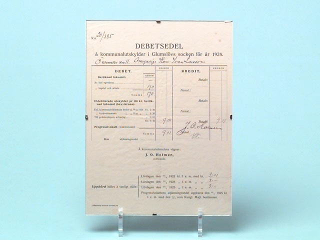 En debetsedel som är inramad i en modern glasram. På debetsedeln står det bl.a: Debetsedel å kommunalutskylder i Glumslövs socken för år 1924. Glumslöv N:o 11. Omyndige Karl Ivan Larsson. Debetsedeln satt på väggen i Ivan Larssons arbetsrum i Borstahusen.