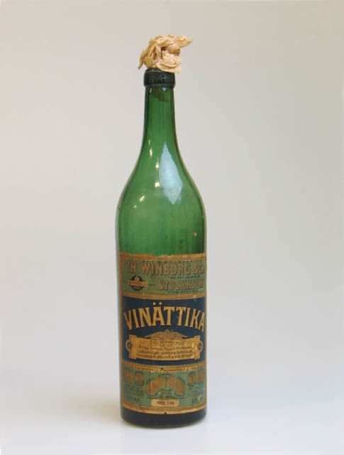 Grön flaska för vinättika. Text på fastklistrad etikett: AKTIEBOLAGET TH. WINBORG & Co, KGL. HOV-LEVERANTÖR, STOCKHOLM, ETABLERAD 1861, VINÄTTIKA, PRIS 1:85.