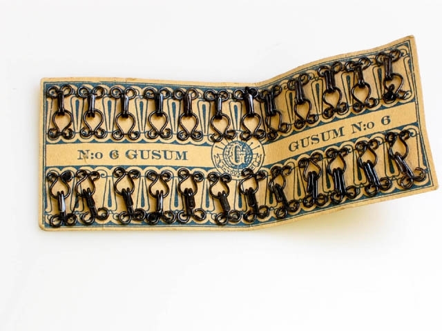 En kartongbit med hyskor och hakar som är fastsydda med små stygn. Text på pappret: "N:o 6 GUSUM, SKYDDSMÄRKE".

De är tillverkade på Gusums Bruk som fanns mellan 1653 - 1988.
År 1707 startar man ett knappmanufakturi med tillverkning av knappar, spännen och andra småartiklar i mässing.
1949 upphör tillverkningen av knappar, spännen, häktor, byglar m.m.