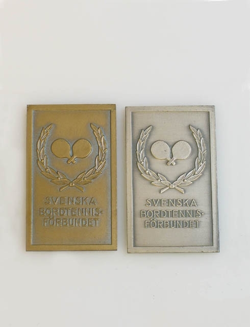 Nio plaketter och tre medaljer från "SVENSKA BORDTENNISFÖRBUNDET". Bild på är två korsade bordtennisracket omringade av lagerblad. Sträcker sig från 1949 till 1963. Två tillverkade av patinerad brons, sju troligtvis av tenn. Stämplad på baksidan: "Lagerströms Mjölby". Medaljerna är upphängda i grönt kort band. Alla med text på baksidan: "Div I 1962-63".
Svenska Bordtennisförbundet skapades 1926.