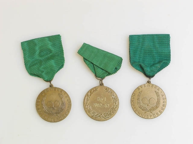 Nio plaketter och tre medaljer från "SVENSKA BORDTENNISFÖRBUNDET". Bild på är två korsade bordtennisracket omringade av lagerblad. Sträcker sig från 1949 till 1963. Två tillverkade av patinerad brons, sju troligtvis av tenn. Stämplad på baksidan: "Lagerströms Mjölby". Medaljerna är upphängda i grönt kort band. Alla med text på baksidan: "Div I 1962-63".
Svenska Bordtennisförbundet skapades 1926.