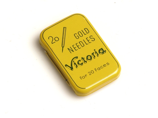Plåtask, gul med mörkgrön bård och texten "20 gold needles Victoria For 20 Faces" och en teckning av en sådan nål som finns i asken. Nålarna är till en automatväxlande skivspelare, enligt guldtexten på det svarta skyddspappret som omsluter nålarna. Det finns 16 nålar kvar i asken. På askens långsida står texten "Registered Model" och på askens kortsida står det "Swiss Made".