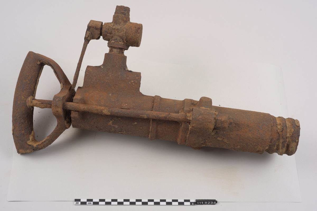 Maskin av metall. Rusten. Sylindrisk utforming. Firkantet hull i ene enden. Håndtak i metall i den andre. Utvendige gjenger i metallet i enden hvor hullet er. Ventil med kran for trykkluftstilkobling på ene siden. Støtdempere løper langs maskinen på to sider.