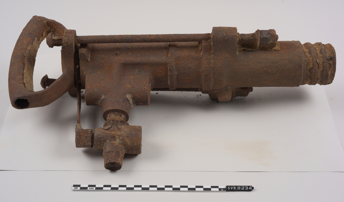 Maskin av metall. Rusten. Sylindrisk utforming. Firkantet hull i ene enden. Håndtak i metall i den andre. Utvendige gjenger i metallet i enden hvor hullet er. Ventil med kran for trykkluftstilkobling på ene siden. Støtdempere løper langs maskinen på to sider.