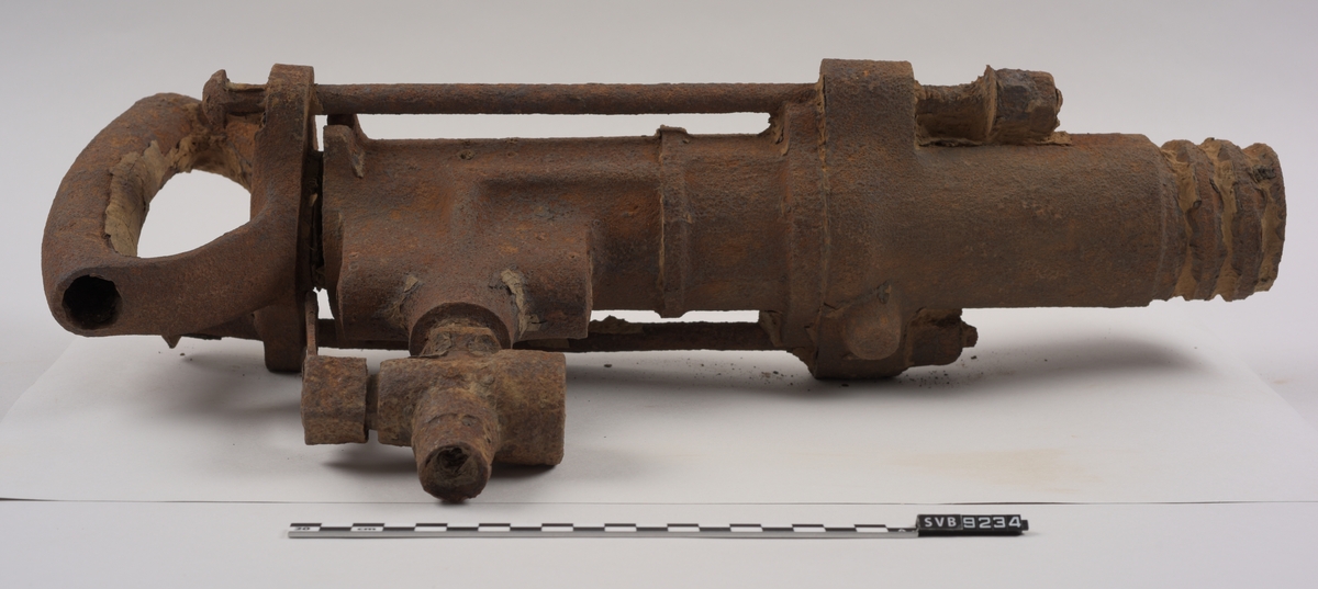 Maskin av metall. Rusten. Sylindrisk utforming. Firkantet hull i ene enden. Håndtak i metall i den andre. Utvendige gjenger i metallet i enden hvor hullet er. Ventil med kran for trykkluftstilkobling på ene siden. Støtdempere løper langs maskinen på to sider.