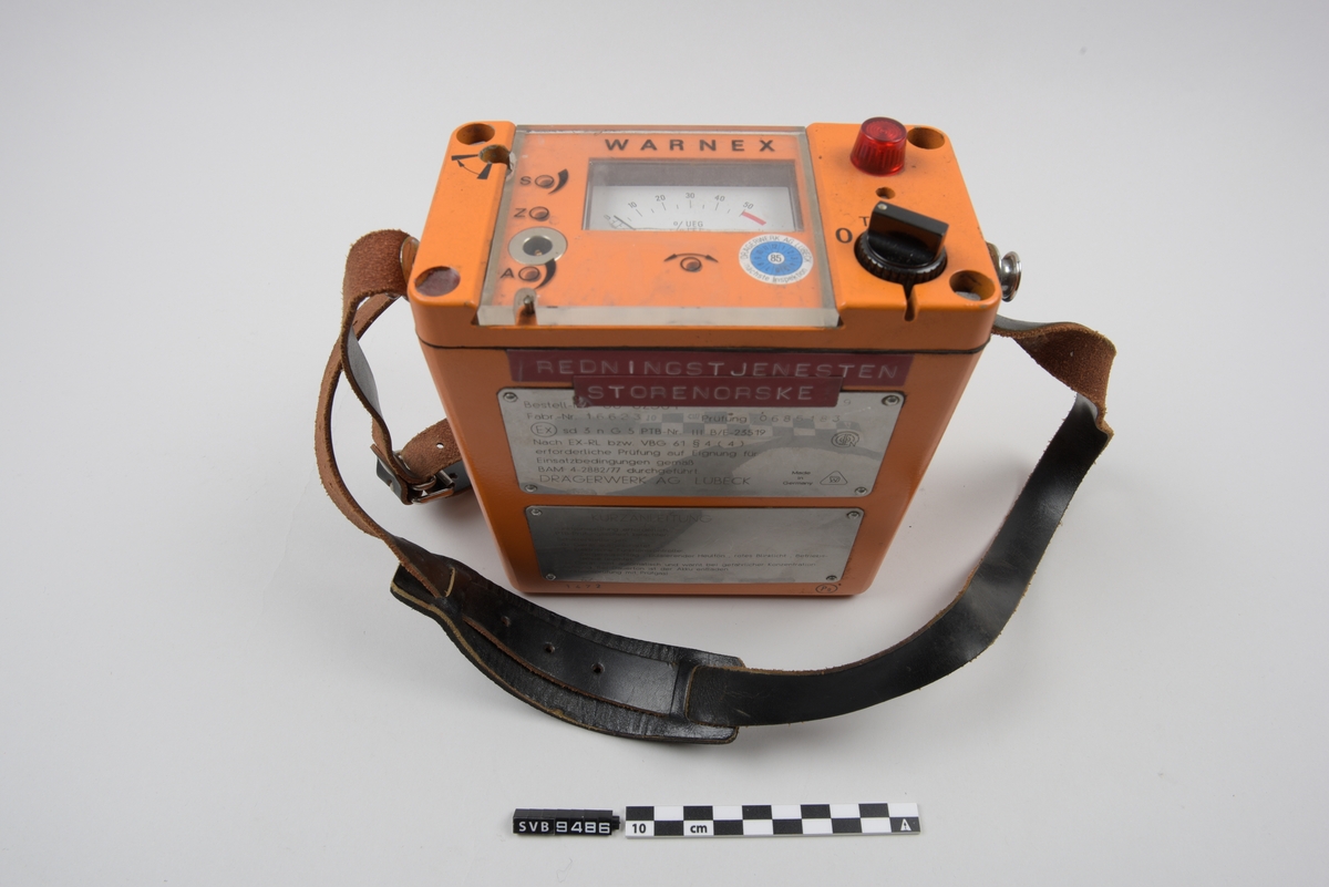 Måleinstrument i oransje metall med sort reim.  På toppen er det en vendebryter, en rød lampe og et panel med gjennomsiktig plast med et dreiespoleinstrument. På venstre side av instrumentet er det et sirkulært plastdeksel med en ventil i midten. Et lignende deksel finnes også på baksiden av instrumentet. I midten av dette er det et sølvfarget stoff. På fremsiden av instrumentet er det to rødeklistermerker og to metallplansjer med tekst. På bunn er det fire gummiknotter.