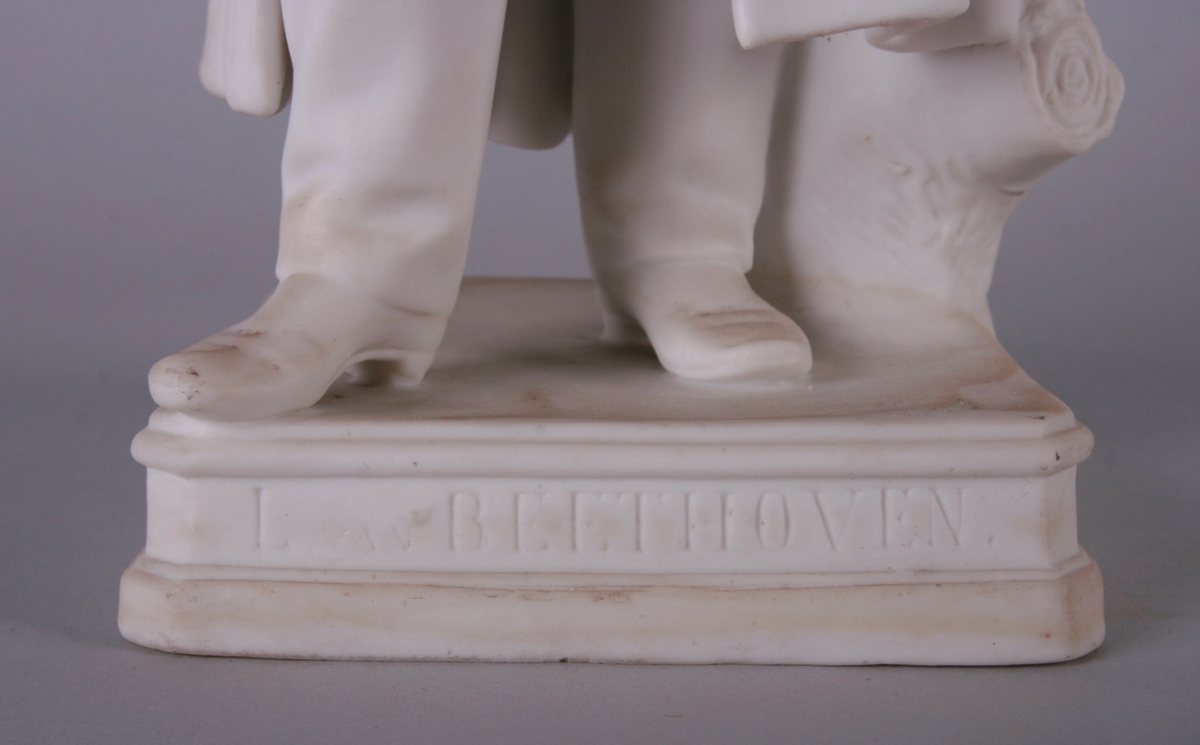 Beethoven stående iført sid, åpen frakk. Tyngden på venstre ben, armene overkors. I høyre hånd holder han en penn, i venstre noen noteark. Figuren står på en kvadratisk plate.