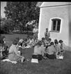 Kvinnliga scoutledare sitter med anteckningsblock i en ring vända mot varandra på en gräsmatta utanför gamla tingshuset på Visingsö. Bakom dem står två manliga scoutledare.