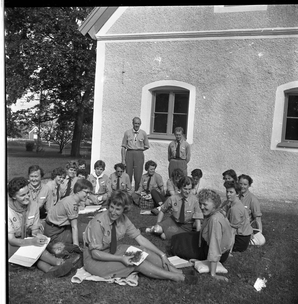 Kvinnliga scoutledare sitter med anteckningsblock i en ring på en gräsmatta utanför gamla tingshuset på Visingsö, de vänder sig mot fotografen. Bakom dem står två manliga scoutledare.