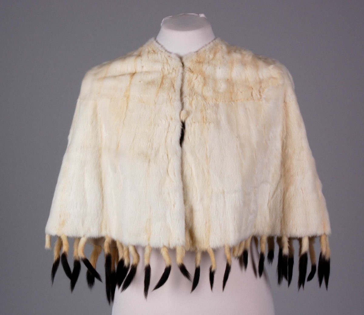 Midjelang cape, med rund, tettsluttende halsringning og lukning med to hekter midt foran. Pelsstykkene er sammensydd slik at det dannes vertikale, smale felt, og de rette skuldrene er formet ved skuldersømmer. Capen er kantet med tette frynser av røyskatthaler i nederkant og helföret med benhvit silke. På innsiden, nede i front, en tollplombering.