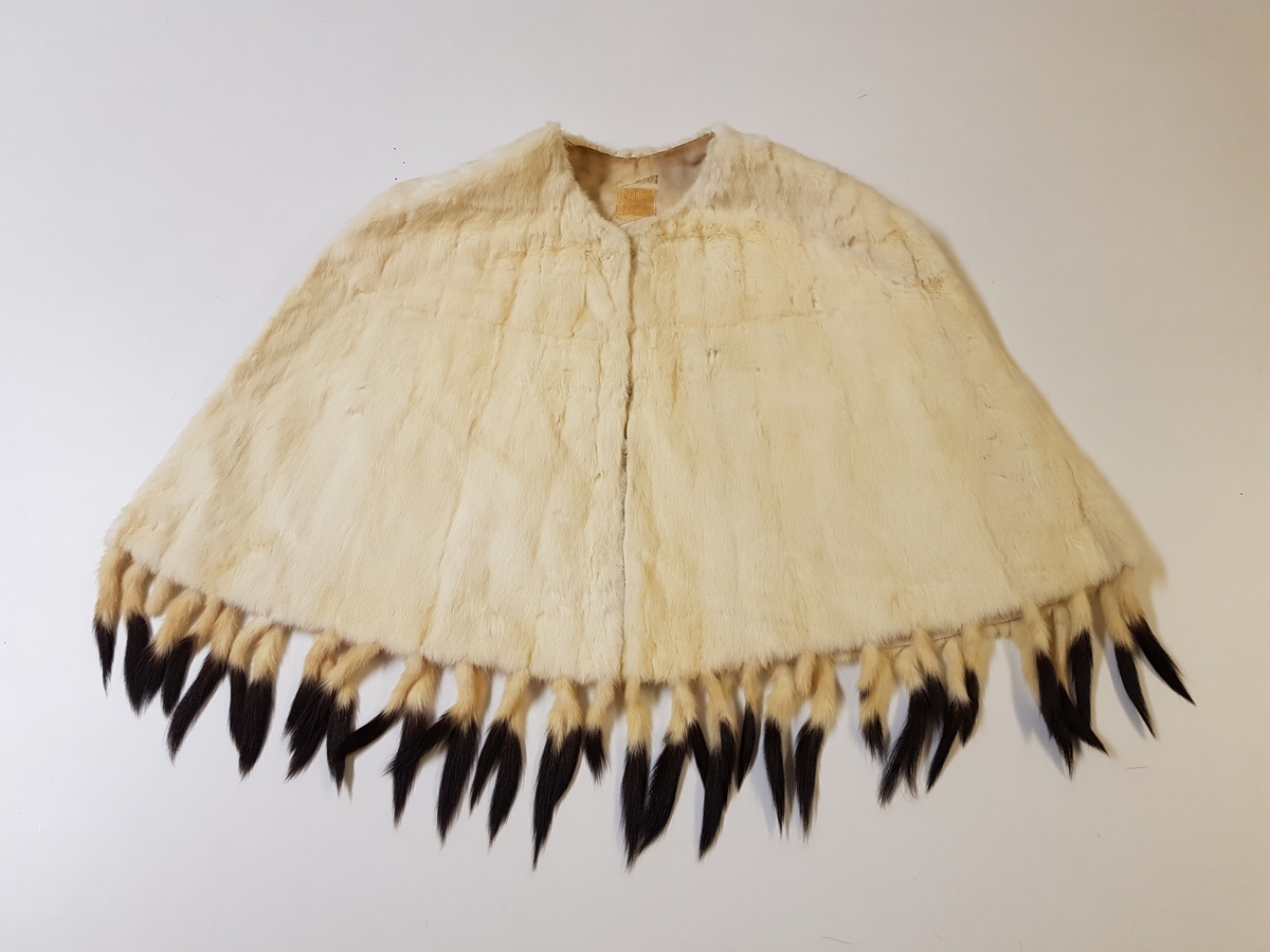 Midjelang cape, med rund, tettsluttende halsringning og lukning med to hekter midt foran. Pelsstykkene er sammensydd slik at det dannes vertikale, smale felt, og de rette skuldrene er formet ved skuldersømmer. Capen er kantet med tette frynser av røyskatthaler i nederkant og helföret med benhvit silke. På innsiden, nede i front, en tollplombering.