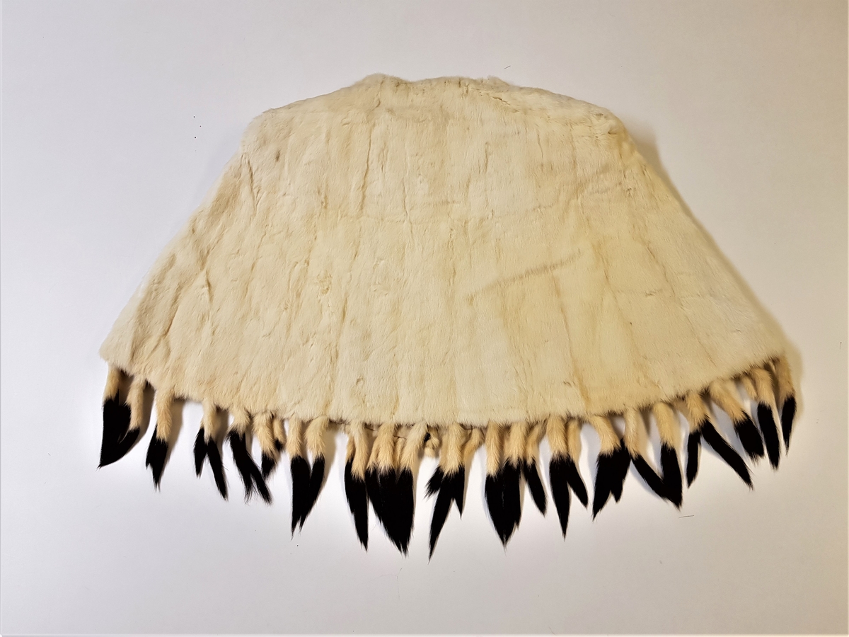 Midjelang cape, med rund, tettsluttende halsringning og lukning med to hekter midt foran. Pelsstykkene er sammensydd slik at det dannes vertikale, smale felt, og de rette skuldrene er formet ved skuldersømmer. Capen er kantet med tette frynser av røyskatthaler i nederkant og helföret med benhvit silke. På innsiden, nede i front, en tollplombering.