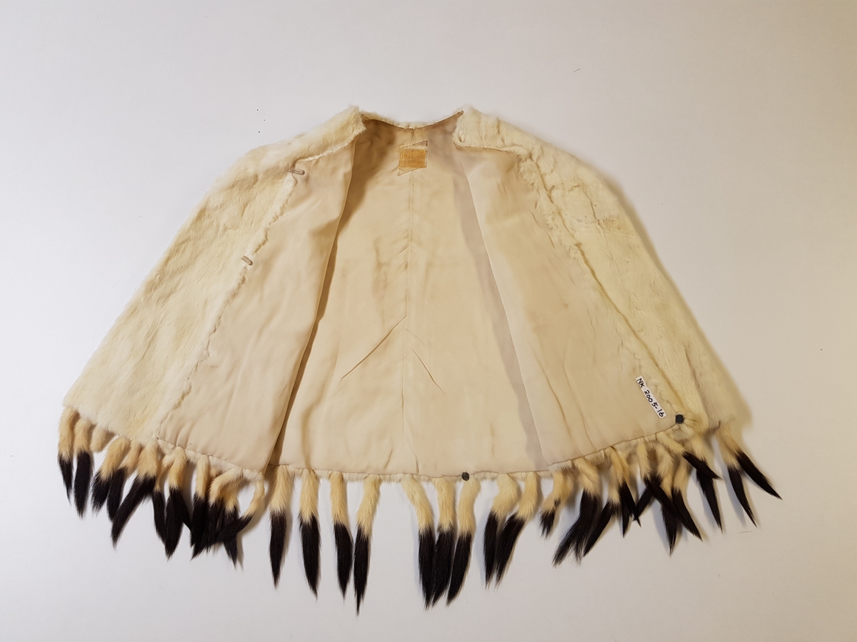 Midjelang cape, med rund, tettsluttende halsringning og lukning med to hekter midt foran. Pelsstykkene er sammensydd slik at det dannes vertikale, smale felt, og de rette skuldrene er formet ved skuldersømmer. Capen er kantet med tette frynser av røyskatthaler i nederkant og helföret med benhvit silke. På innsiden, nede i front, en tollplombering.