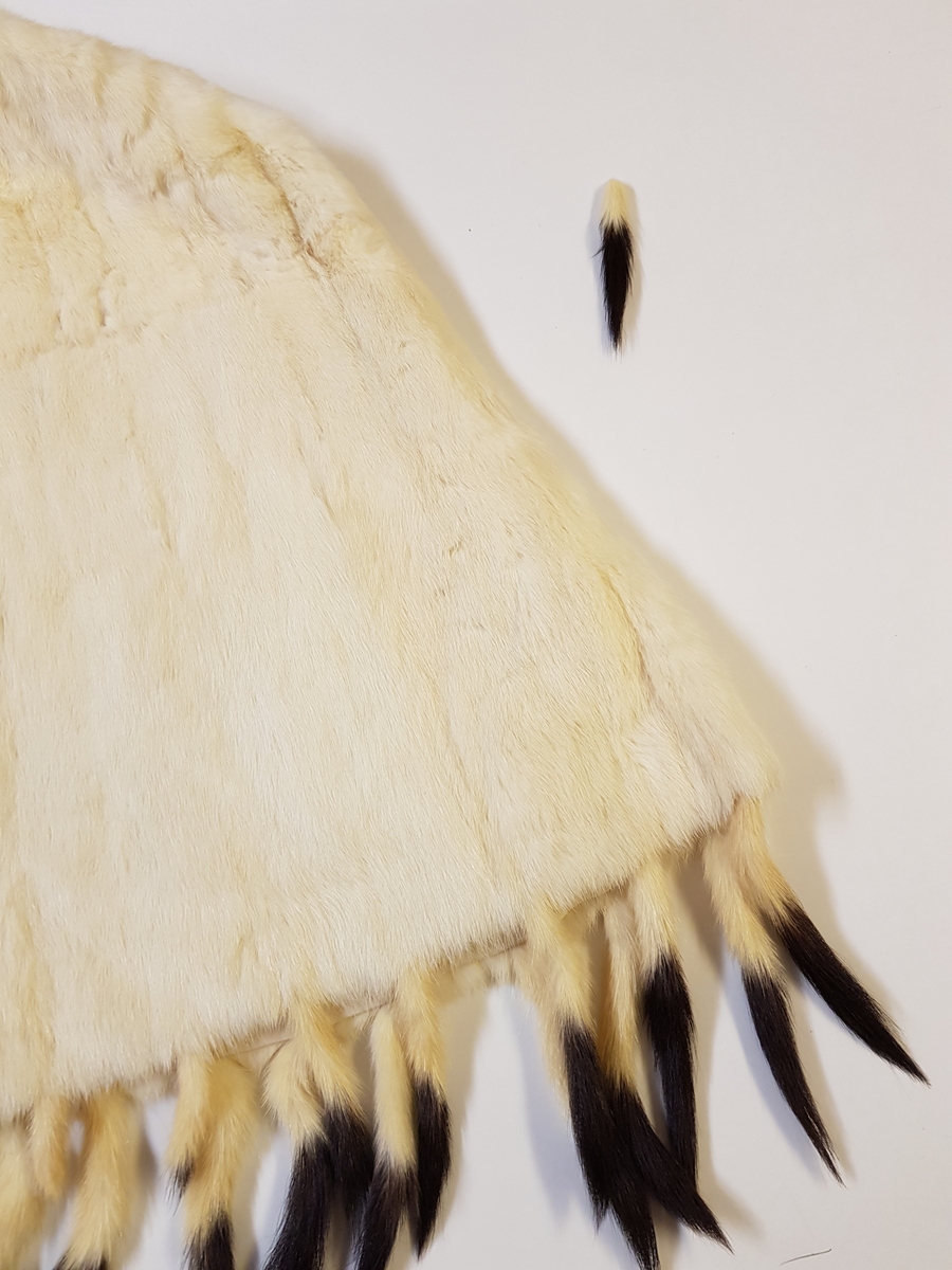 Midjelang cape, med rund, tettsluttende halsringning og lukning med to hekter midt foran. Pelsstykkene er sammensydd slik at det dannes vertikale, smale felt, og de rette skuldrene er formet ved skuldersømmer. Capen er kantet med tette frynser av røyskatthaler i nederkant og helföret med benhvit silke. På innsiden, nede i front, en tollplombering.