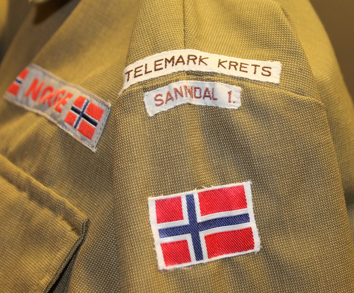 Sid skjorte med lang erme og brystlomme. A-formet skjørt i 5 bredder. To utenpåsydd lommer med klaff og knapp. Lukking bag med glidelås og knapp. Påsydd merkelapper med navn og flagg og ei gull stjerne (5 års tjeneste).
Over 
Tilbehør:: grønn og rød snor og 5 sjerf.