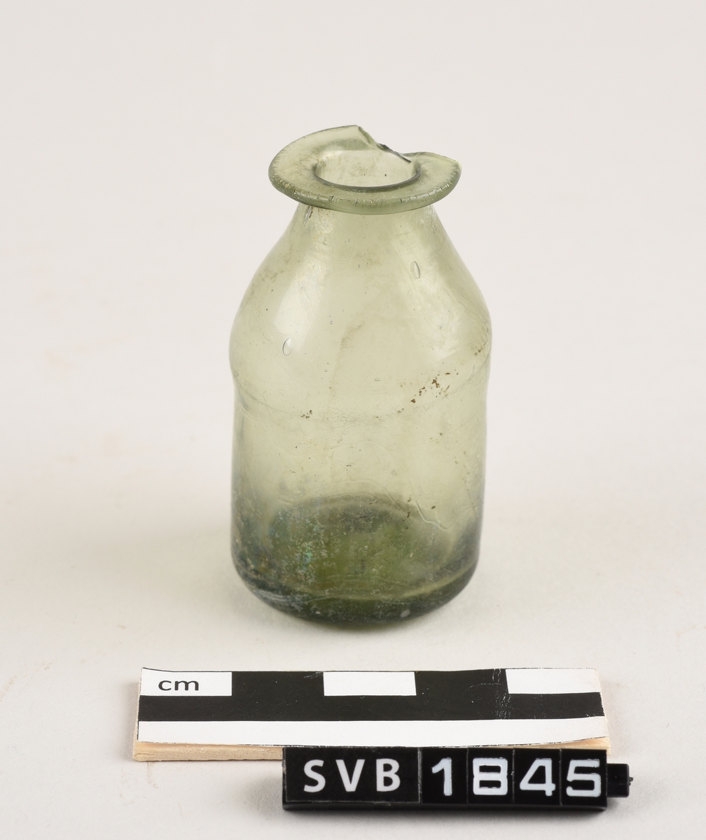 Sylindriskformet glassflaske. Deler av randen mangler. På toppen av flasken er det et sirkelformet hull. Hullet har en diameter på 1,2 cm. Flasken er lys grønn i farge.