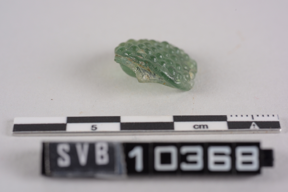 Fragment av glass. Grønn farge. Det er dekor på den ene siden. Dekoren er som små konvekse halvkuler.