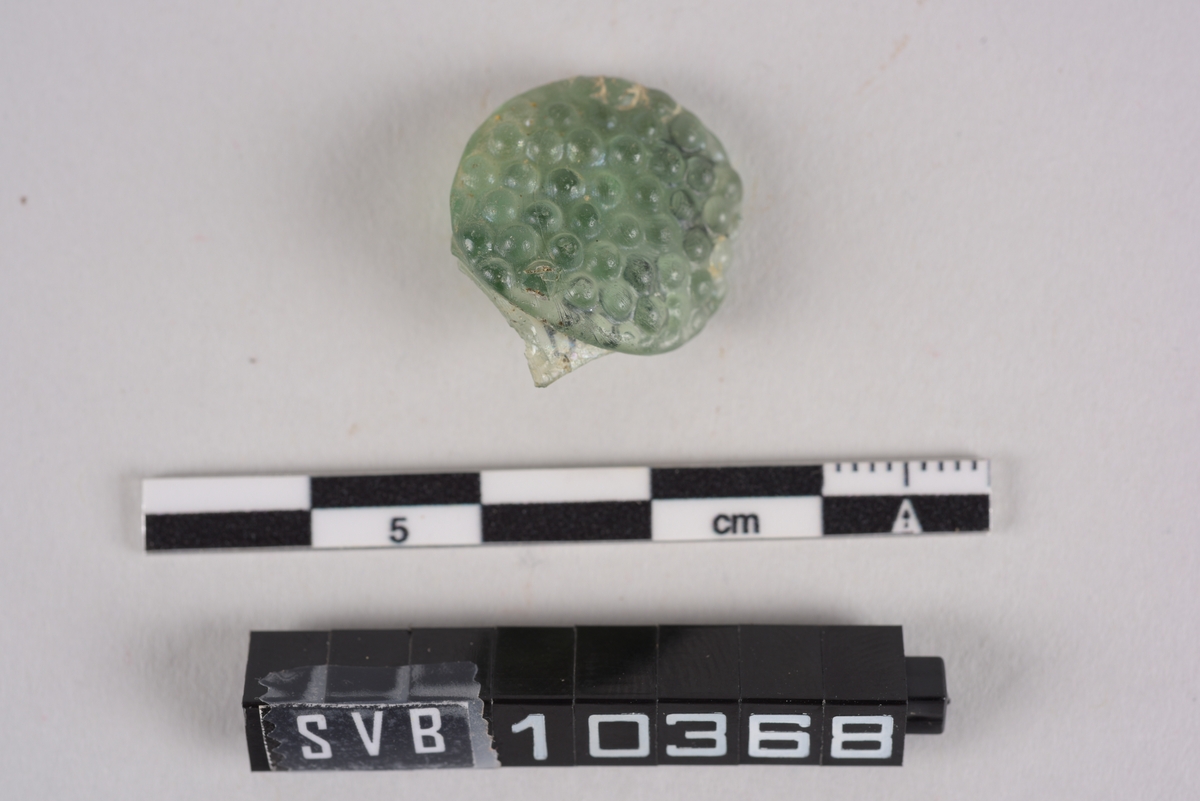 Fragment av glass. Grønn farge. Det er dekor på den ene siden. Dekoren er som små konvekse halvkuler.