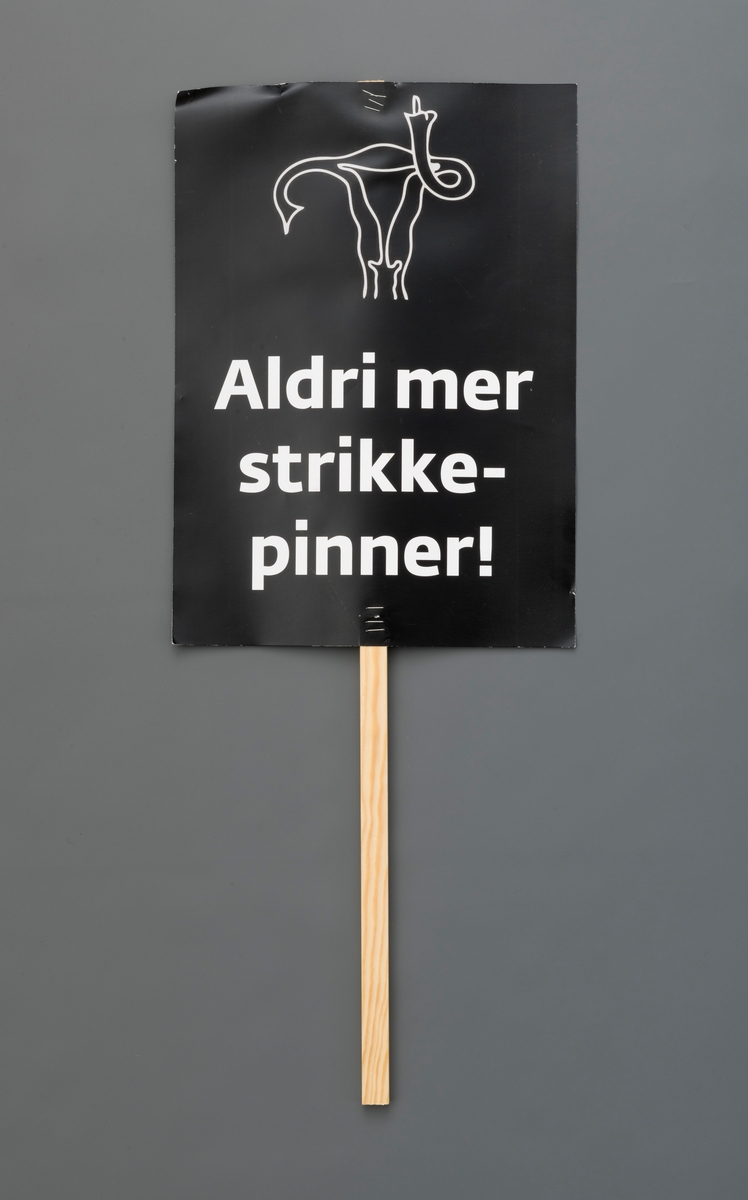 Ferdig trykket plakat stiftet på trepinne/-håndtak. Sort bunn med hvit tegning av livmor som viser fingeren og hvit tekst: "Aldri mer strikkepinner!"