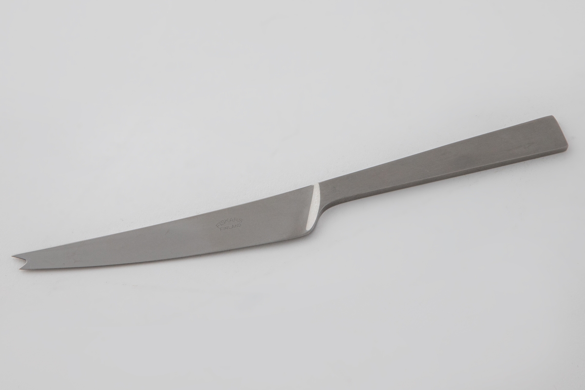 Ostekniv i polert, rustfritt stål. Blad med svakt kurvet rygg, som ender i en gaffelformet avsluttning. Det rettlinjede skaftet er mattert, mens bladet er polert, slik at det oppstår en kontrast mellom de blanke og matte partiene.