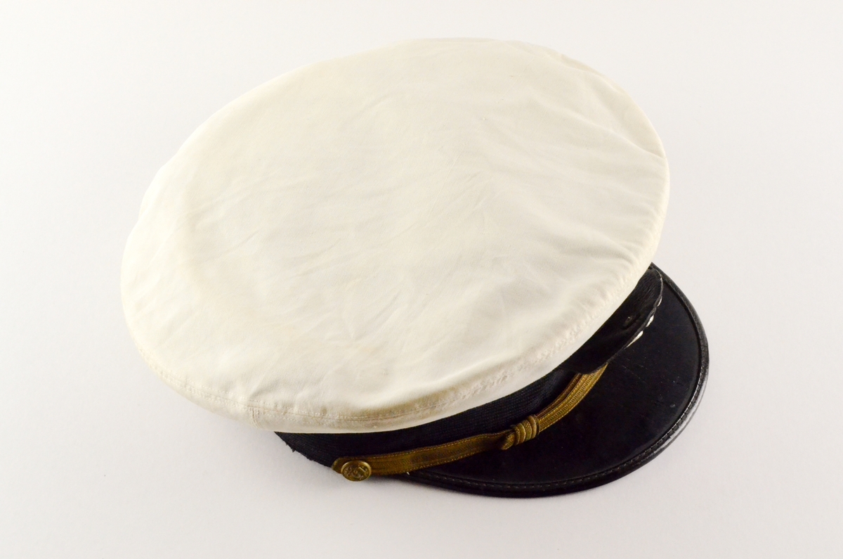 Uniformslue med skygge. Hvit rund flat pull, sort hattebånd med påsatt gulltråd, tynnere i front. Midt foran på pull og bånd: emblem i metall; sølvfarget ørn på lfagg, gullfargede anker. Sort skygge. Kanten stivet innvendig med brunt lærbånd.