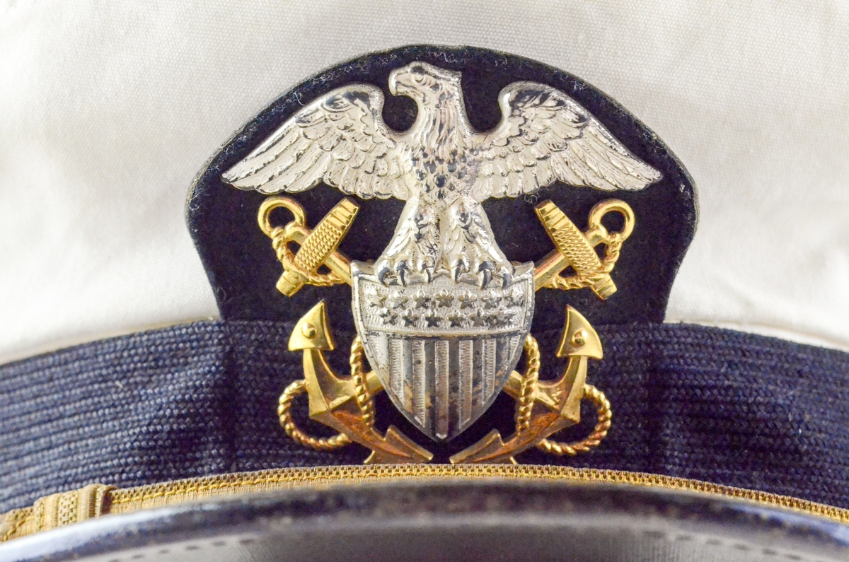 Uniformslue med skygge. Hvit rund flat pull, sort hattebånd med påsatt gulltråd, tynnere i front. Midt foran på pull og bånd: emblem i metall; sølvfarget ørn på lfagg, gullfargede anker. Sort skygge. Kanten stivet innvendig med brunt lærbånd.