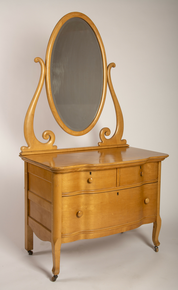 Kommode. Topplate og fronten på kommoden er bølget i formen. To mindre skuffer øverst, og en stor låsbar skuff nederst. Treknotter. Ben med hjul.