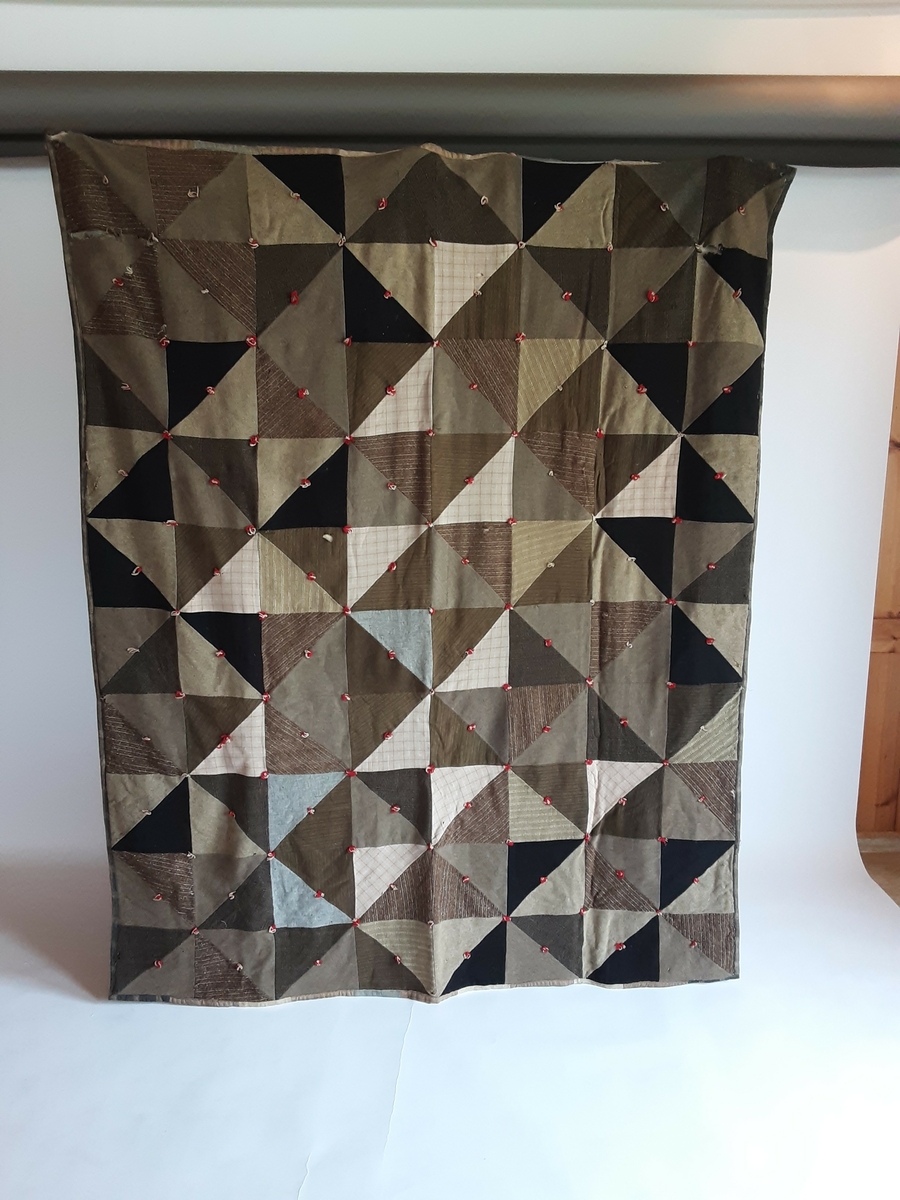 Vatt-teppe. Quiltet, brune nyanser. Satt sammen av trekanter. Ulike litt grove stofftyper (ull?). Pyntet med knyter laget av rødt og beige garn. De samme stoffene er satt sammen til striper og danner baksiden på teppet. Huller i stoffet enkelte steder.