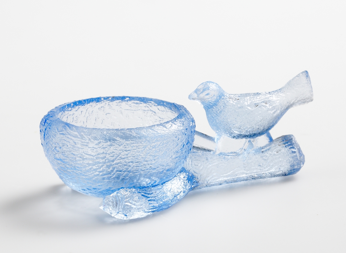 Saltkar eller äggkopp, enligt katalogen som förmedlades i Lindshammars glasbruk (se bifogad bild), i blåtonat glas, i form av ett fågelbo och en fågel på en grenklyka.