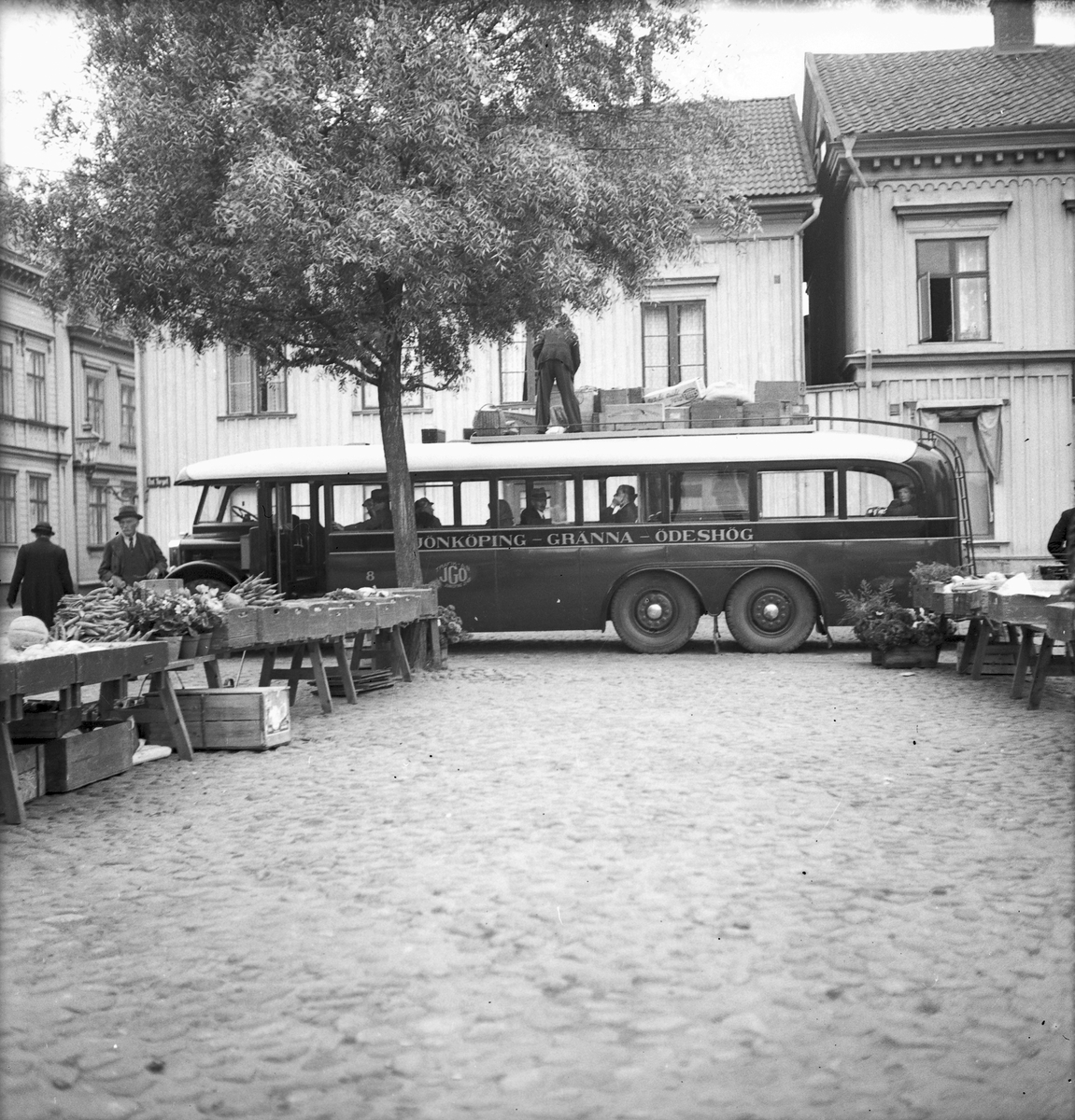 På Östra Torget i Jönköping handlas det framför allt med grönsaker. Bussen på linjen Jönköping-Gränna-Ödeshög har stannat till och busschauffören står på taket och surrar bagage. Till vänster i bild, söder om torget ligger Slottsgatan.