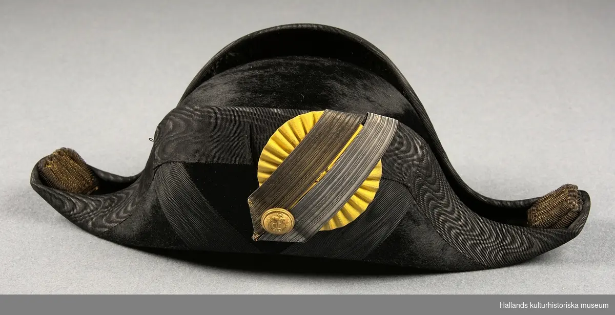 A: Hattask av papp målad i en lilablå nyans. På ovansidan ett emblem från tillverkaren och på baksidan ett snöre.

B: Befällshatt av bicornetyp. Hatten är klädd med svart fälb och ett svart mönstrat sidenband. På höger sida sitter en gul sidenkokard under dubbla band - ett i guld och ett i silver. På höger sida även en guldknapp med flottans vapen bestående av ett ankare och tre kronor. Guldfärgade tassar bak- och framtill på hatten. Hakrem av svart läder.