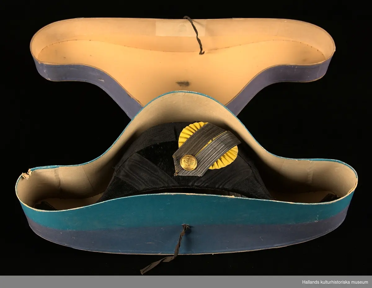 A: Hattask av papp målad i en lilablå nyans. På ovansidan ett emblem från tillverkaren och på baksidan ett snöre.

B: Befällshatt av bicornetyp. Hatten är klädd med svart fälb och ett svart mönstrat sidenband. På höger sida sitter en gul sidenkokard under dubbla band - ett i guld och ett i silver. På höger sida även en guldknapp med flottans vapen bestående av ett ankare och tre kronor. Guldfärgade tassar bak- och framtill på hatten. Hakrem av svart läder.