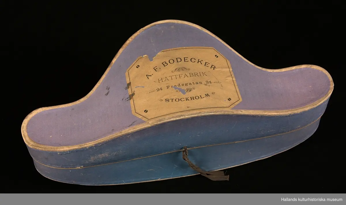 A: Hattask av papp målad i en lilablå nyans. På ovansidan ett emblem från tillverkaren och på baksidan ett snöre.

B: Befällshatt av bicornetyp. Hatten är klädd med svart fälb och ett svart mönstrat sidenband. På höger sida sitter en gul sidenkokard under dubbla band - ett i guld och ett i silver. På höger sida även en guldknapp med flottans vapen bestående av ett ankare och tre kronor. Guldfärgade tassar bak- och framtill på hatten. Hakrem av svart läder.