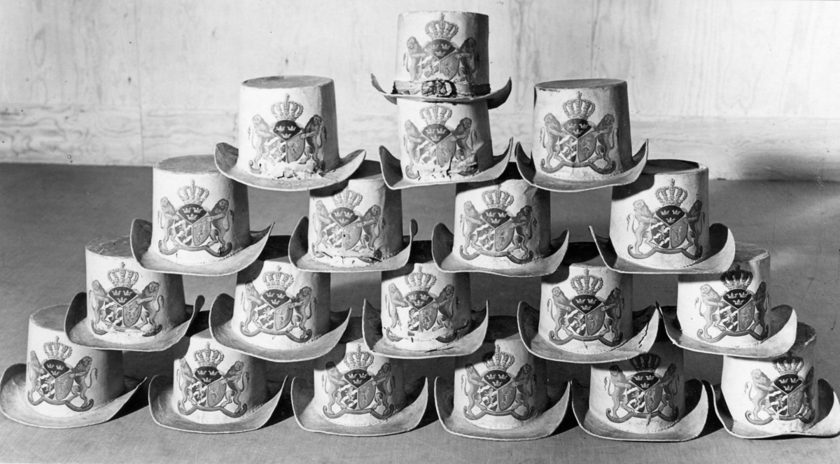 Hatt tillverkad av papp som målats med en crémegul färg. Framtill försedd med svensk-norska riksvapnet i färg. Foder av bomullstyg, 115 mm brett och dragsko.