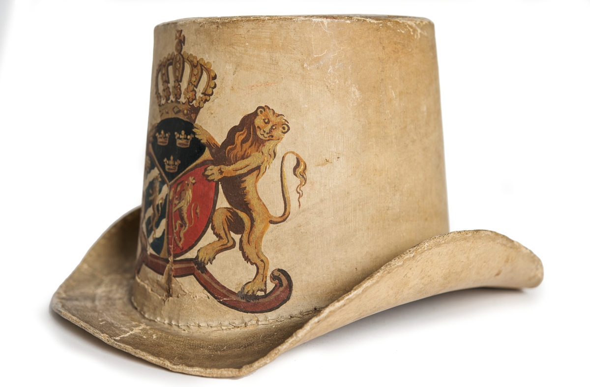 Hatt tillverkad av papp som målats med en crémegul färg. Framtill försedd med svensk-norska riksvapnet i färg. Foder av bomullstyg, 115 mm brett och dragsko.