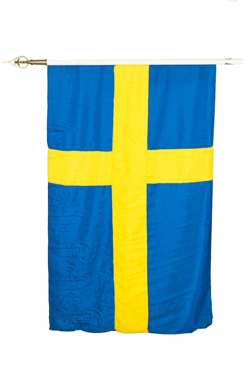 Svensk flagga i siden på vitmålad trästång. Stången har mässingsbeslag och knopp av mässing föreställande en Vasakärve. Tillhörande fodral av grågrönt bomull och läder finns.