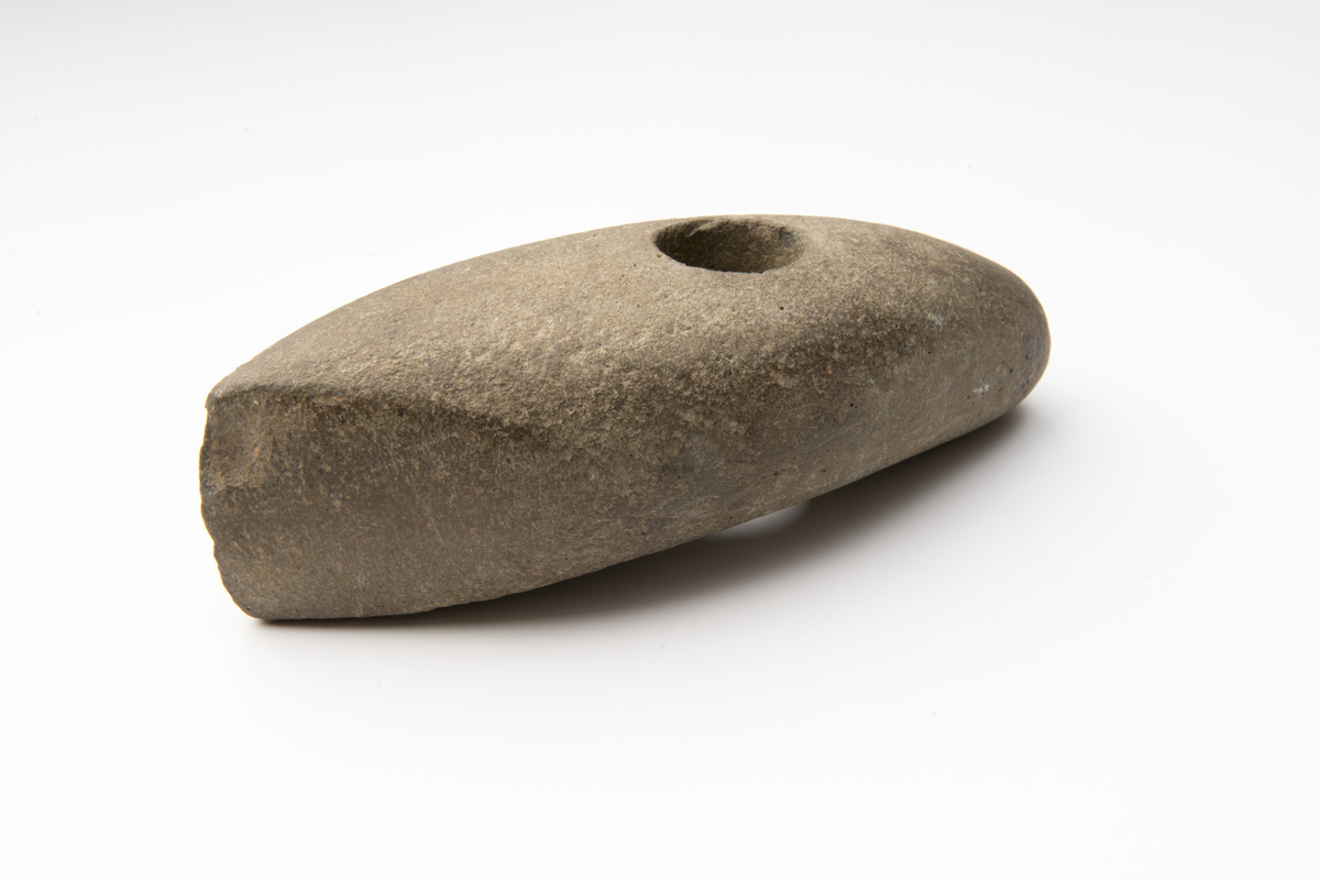 "Vakkert eksemplar av skafthulløks av gabbro. Minner en del om Madsen: Danske Oldsager og Mindesmerker fra Stenalderen, s. 32 fig. 23. Eggog nakkeutformingen er lik, men vårt eksemplar er omtrent dobbelt så langt, og eggslipingen når bare halvveis fra odden til skafthullet. Nakkepartiet er bøyd, og i sidesnitt er øksa mer krum enn typeeksemplaret. Lengde 19,3 cm, største bredde 8,5 cm (fig. 9)." I samsvar med beskrivelsen.
