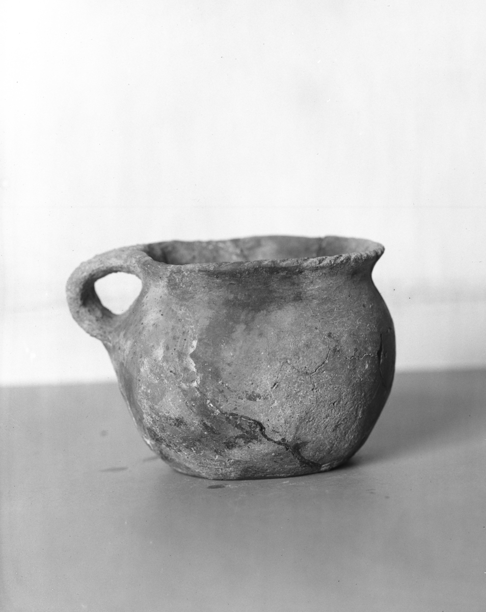 "2397 b. Urnekop, lig N. O. 364, men uden ornamenter. Kanten er forsiret med skraafurer. Den er 9,8 cm høi, 11,9 cm vid over aabningen og 7,5 cm vid over bunden. " Ny beskrivelse etter revisjon:   Hankekar som R364 uten dekor, avb. Bøe fig. 256.  Karet er limt sammen og delvis 'restaurert'.  Rand diam: 117-120 mm, buk diam: 125-128 mm. Randen er dekorert med neglehakk (snorimitasjon).